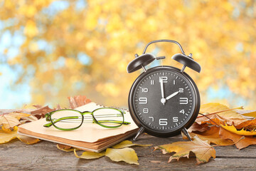 Alarm clock, notebook, glasses and autumn leaves on table outdoors