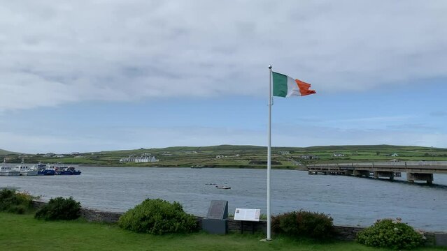Irish flag on a big white pole is waiving from a heavy wind. View from Portmagee city park. Valentia island and fishing, tourist boats in the background. Motion of water. Portmagee, Kerry, Ireland