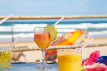 Drinks tropicais em mesa de praia no nordeste do Brasil. No fundo ondas do mar e um guarda sol colorido