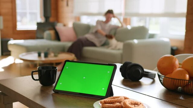 Tablet computer with green screen chromakey display with motion trackers laying on table near oranges, cookies and cup of hot tea. woman playing with dog on cozy sofa in living room on background