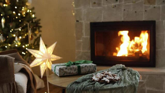 Cozy christmas eve. Stylish wrapped gift and gingerbread cookies on table against festive decorated christmas tree with golden lights and burning fireplace. Winter hygge footage