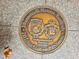 Bronzetafel, die in einem Gehweg eingebettet ist. Die Tafel ist auf Deutsch und lautet “20 Jahre St. Nazaire Saarlouis Städtepartnerschaft Jumelage 1969-1989”. 