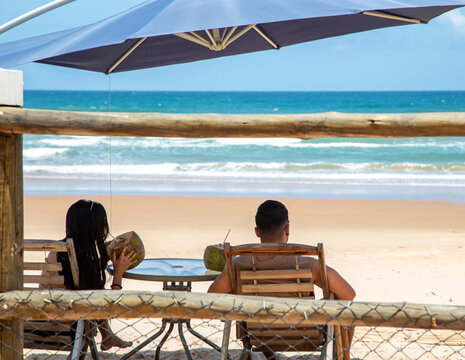 Jovem casal tomando &aacute;gua de coco na sombra em um resort em uma praia do nordeste brasileiro