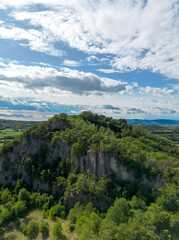 Obraz premium Aerial view of steep cliff on a hill with green trees growing on top below blue sky with clouds in Italian countryside landscape