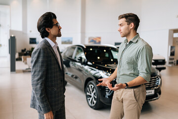 Side view of handsome male client talking to car dealer wearing business suit in dealership discussing automobiles looking at luxurious new model. Concept of choosing and buying new auto at showroom.