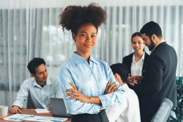 Young African businesswoman poses confidently with diverse coworkers in busy meeting room background. Multicultural team works together for business success. Office lady portrait. Concord