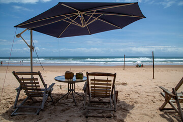 Cocos com canudinho na mesa e cadeiras de praia vazias esperando turistas em praia ensolarada do nordeste brasileiro, com mar e ondas no horizonte