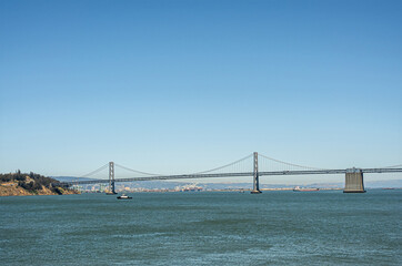 Fototapeta premium San Francisco, CA, USA - July 13, 2023: SF-Oakland Bay Bridge lands on Yerba Buena Island under blue sky above blue water. Ships on horizon