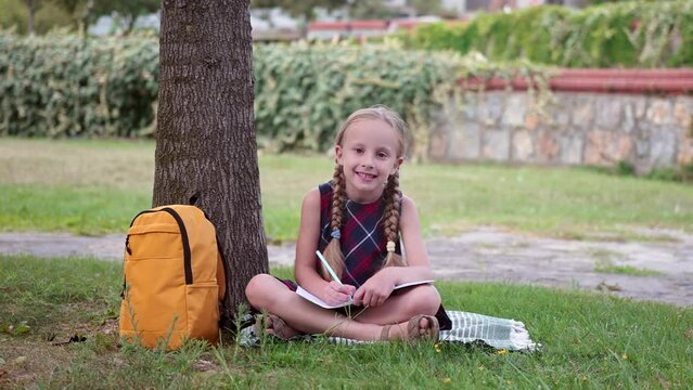 Smiling little girl in dress does homework sitting on grass in school yard happy schoolgirl writes in exercise book resting in park elementary school student