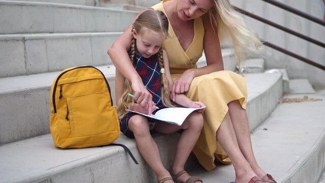 Careful mother helps little daughter with homework sitting on stone step seat at stadium woman with schoolgirl do learning tasks on city street happy family