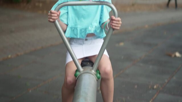 Smiling little girl in blouse with ruffles plays seesaw in summer urban park happy child rides attraction on children playground schoolgirl has fun in city garden