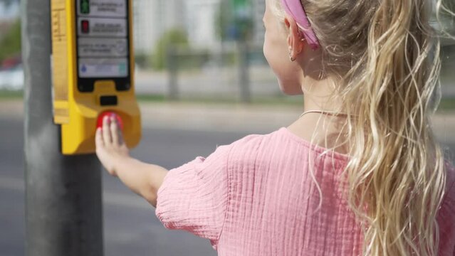 Little girl with blonde ponytail pushes cross street button standing on sidewalk closeup schoolgirl presses key to control streetlight in city child and traffic safety