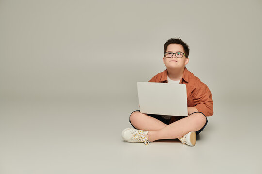 stylish and thoughtful boy with down syndrome sitting with laptop and crossed legs on grey - Powered by Adobe