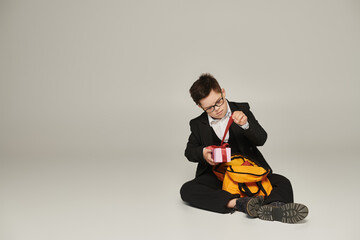 schoolboy with down syndrome, in black uniform sitting near backpack and opening gift box on grey