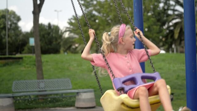Blonde little girl rides swings in green city park cute child in pink dress enjoys swinging on children playground in garden kid plays attraction on gloomy day