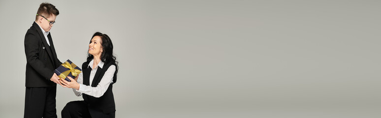 smiling woman in formal wear presenting gift to son with down syndrome on grey, banner