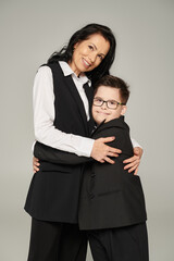 cheerful schoolboy with down syndrome embracing mother in formal wear and looking at camera on grey