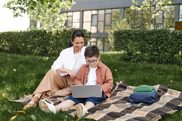 boy with down syndrome, in eyeglasses, sitting with laptop in park near happy mother, e-learning