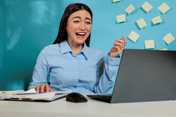Optimistic asian woman having a funny video call conversation sitting at office desk. Female company employee laughing with excitement while talking to colleague remotely.