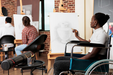 Disabled African American girl in wheelchair drawing on canvas during group art class, improving...