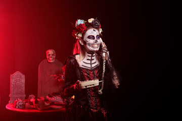 Spooky woman answering landline phone with cord, talking on office telephone while she has skull make up and body art. Portraying santa muerte lady of death on day of the dead celebration.