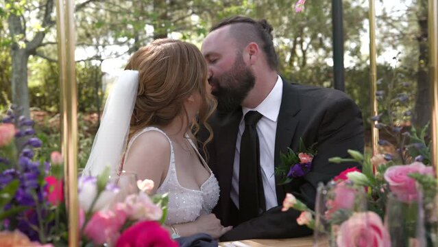 Fair-haired bride and middle-aged groom with black beard sitting at wedding table, decorated with colorful bouquets, kissing, smiling. Outdoor ceremony. High quality 4k footage