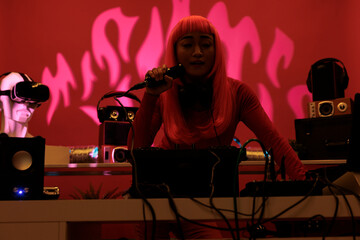 Cheeful artist with pink hair standing at dj table playing electronic music with turntables,...