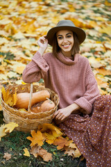 Fashionable woman in cozy knitted sweater, elegant dress, and chic hat sits beside a basket with pumpkins. Surrounded by golden autumn leaves on the ground