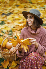 Happy attractive woman in knitted sweater, elegant dress and hat sits beside a basket with pumpkins. Holding in hand maple leaf