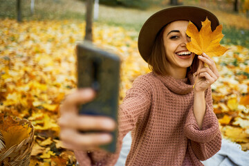 Smiling autumn woman in hat and knit sweater captures the moment with selfie on smartphone at park with maple leafe in hand