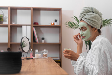 Concentrated woman applying homemade clay mask on face looking in table mirror pretty lady in bathrobe cleansing face with organic cosmetics at home side view