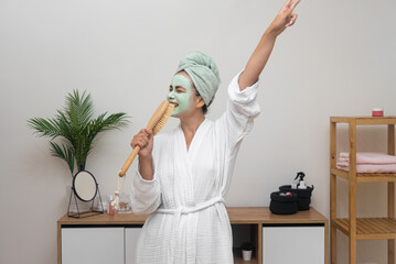 Happy woman with clay mask on face singing in hairbrush mic and dancing at home lady in bathrobe...
