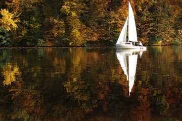 a sailboat and an autumn forest of red yellow and green autumn colors are reflected in the lake surface