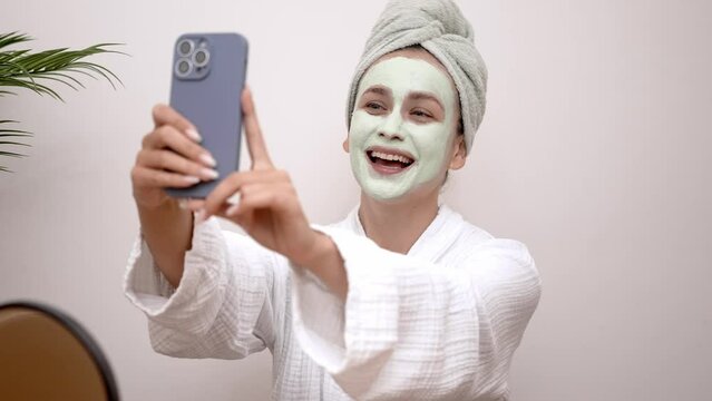 Woman with natural clay mask on face in bathrobe making selfie on smartphone at table lady blogger recording content about skincare for social media in home studio