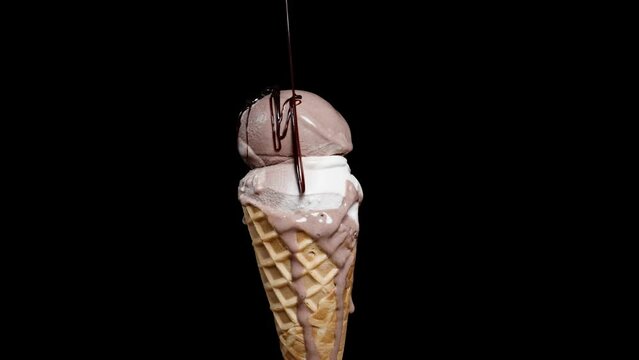 A waffle cone with ice cream scoops spins on a black background, and chocolate is poured over it from above. Isolated on a black background.