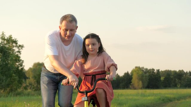Happy father supports bicycle while little girl learning to ride in green field