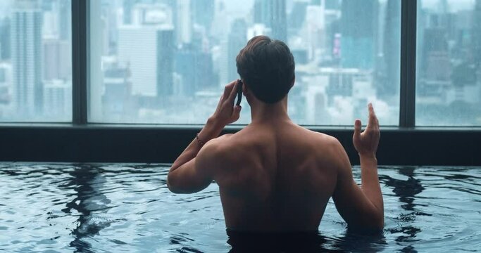 Back view business man talking phone while relax in roof top swimming pool of skyscraper in big city. Rear view businessman uses smartphone for negotiations while relaxing in pool on city background
