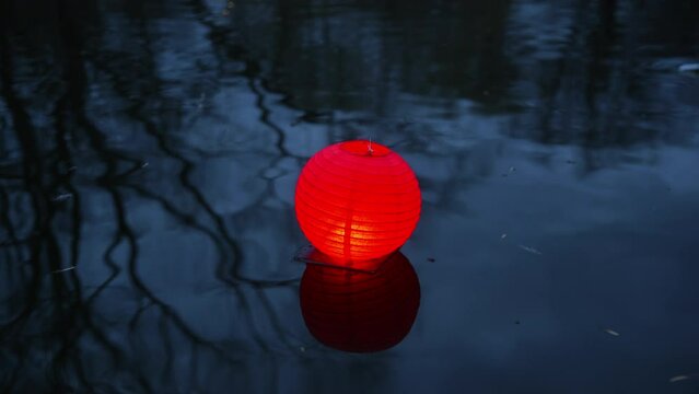 Handheld shot of red illuminated paper lantern floating on river water. Cultural celebration during traditional festival at night. Reflection of bare trees. 