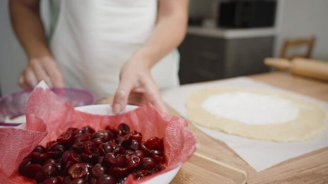 A plate with pitted cherries is in the foreground, which the woman takes and places the cherries onto the dough.
