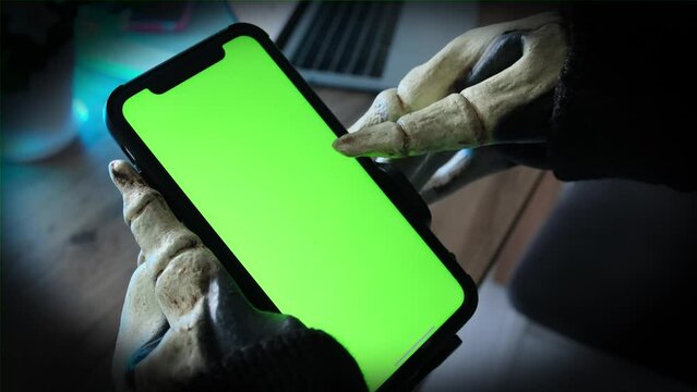 Close up, scary skeletal hand with bony fingers touches the screen with smartphone stroking screen chromakey, dark vignette. Man in skeleton costume becomes obsessed with messaging strangers online