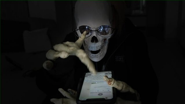 Mysterious man with mask of skull and black hood, poking his bony fingers on smartphone screen in black room, receding tracking shot. Copyspace. Celebrating mystical holiday Halloween.