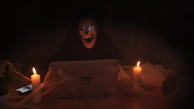 Skeleton in a black hood typing on a laptop sitting in a cobwebby crypt with burning candles in the fog, sending out an invitation to a creepy party, Happy Halloween.