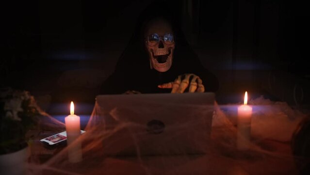 Mystical background old death sitting laptop typing creepy posts on social networks sending out invitations to party in honor Happy Halloween next to burning candles surrounded by cobwebs and fog.