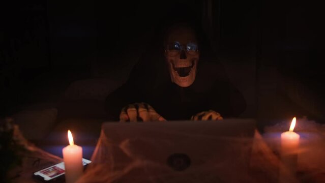 Man in creepy death costume in the misty haze with finger bones and skull mask types at laptop in online chat, Halloween party, making mailing about advertising scary game and creepy posts.