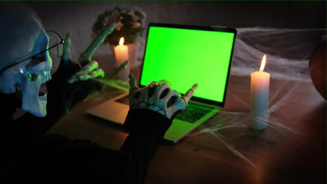 Terribly scary character of death, typing at laptop with green screen and show rock gesture bone hand , side view, close up. Happy Halloween night party masquerade  invitation.