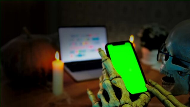 Celebrating holiday, death, bone hand strokes green smartphone screen, terribly spooky Halloween background, dark vignette. Frightening and unforgettable Halloween invitation.