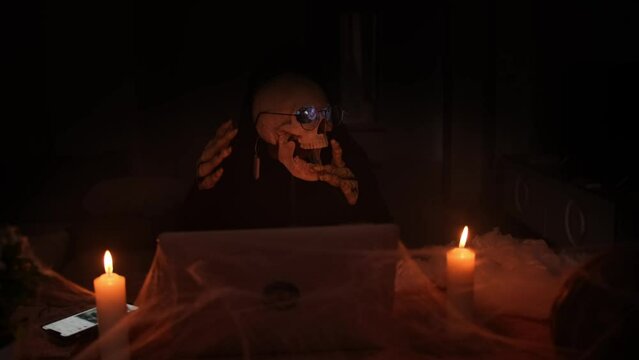 Frightening skeleton costume guardian of hell dancing and typing laptop surrounded by cobwebs and fog, sitting in dark. Celebrating mystical holiday Happy Halloween.