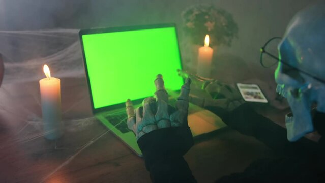 Close up, in fog terribly scary character of death, typing bone hand party invitation at laptop with green screen, side view. Happy Halloween night masquerade. Technology and social media.