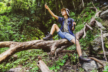Naklejka na ściany i meble Young man, mountain forest explorer caucasian traveler solo adventurer, sitting on a big log, holding compass to find direction, hiking passing through the beautiful waterfall path is satisfying. 