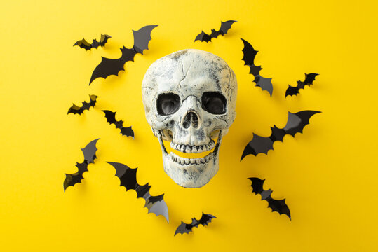Halloween night frights concept. Above view photo of human skull surrounded by black paper bats on yellow isolated background with copy-space for text or advertising